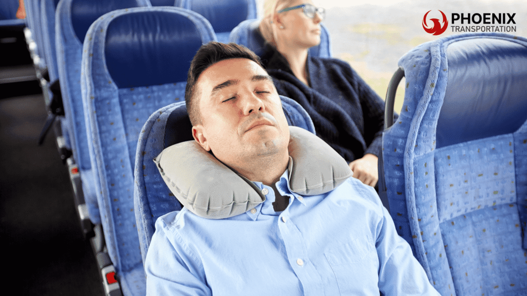 Man sleeping with a neck pillow on a blue seat during a charter bus traveling to Oakland, while a relaxed woman in sunglasses sits behind.